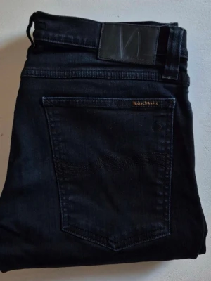  Svarta Nudie Jeans tight long John  - Snygga mörkblå slim fit jeans från Nudie Jeans med klassisk femficksdesign och diskret logga på bakfickan. Jeansen har raka ben och är tillverkade i ett mjukt denimtyg som sitter skönt. Perfekta för en avslappnad och stilren look.