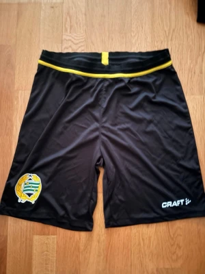 Hammarby shorts  - Säljer ett par svarta sportshorts från Craft med gult band i midjan och klubbmärke på vänster ben. Shortsen är tillverkade i ett lätt och snabbtorkande material, perfekta för träning eller match. Klassisk passform och elastisk midja för skön komfort.