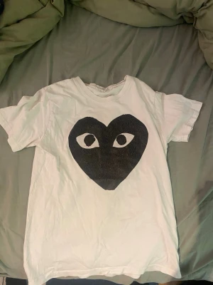 Cdg T-shirt  - Vit t-shirt med ett stort svart hjärta och ögon på bröstet. Klassisk rund hals och korta ärmar. Tillverkad i mjuk bomull, perfekt för en avslappnad och trendig look.
