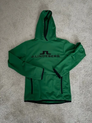 J.Lindeberg hoodie  - Snygg grön hoodie från J.Lindeberg med svart logga och text på bröstet. Hoodien har svarta detaljer längs huva, ärmslut och fickor. Tillverkad i ett slätt, sportigt material som känns modernt och fräscht. Perfekt för dig som vill sticka ut med stilren design.