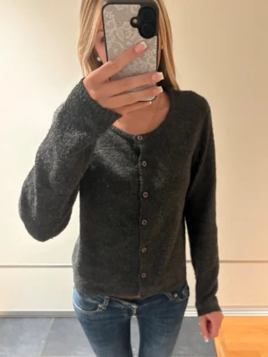 Mörkgrå mysig kofta med knappar - Mysig mörkgrå kofta med rund halsringning och knäppning framtill. Koftan har långa ärmar och en mjuk, lite luddig textur som ger en avslappnad look. Perfekt att slänga över en t-shirt eller linne för extra värme.