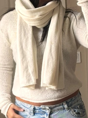 Krämvit mjuk halsduk - En lång och mjuk halsduk i krämvit färg. Halsduken har en enkel, klassisk design utan fransar och är tillverkad i ett fluffigt material som ser riktigt mysigt ut. Perfekt att svepa runt halsen för extra värme under kyliga dagar.