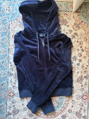 Mörkblå velour hoodie Juicy Couture - Mörkblå hoodie i mjuk velour från Juicy Couture. Klassisk modell med dragkedja, fickor fram och justerbar huva med snören. Diskret Juicy Couture-logga broderad på bröstet. Perfekt för en chill och trendig look.