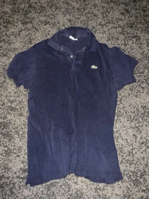 Mörkblå pikétröja med krage - En klassisk mörkblå pikétröja med korta ärmar och krage. Tröjan har knappslå framtill och en liten broderad logga på bröstet. Perfekt till jeans eller shorts för en avslappnad look.