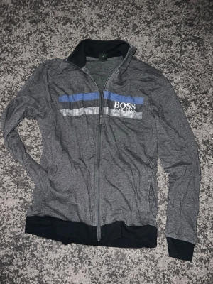 Grå ziptröja från BOSS - Snygg grå ziptröja från BOSS med blå och vita ränder över bröstet och logga. Tröjan har hel dragkedja, svart krage och muddar. Perfekt för en avslappnad men stilren look.