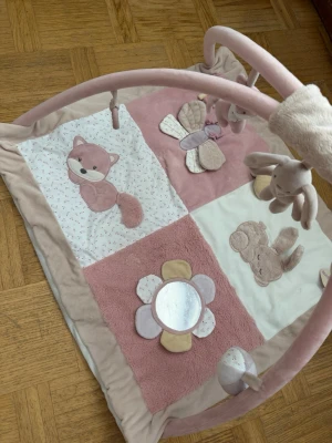 Rosa babygym med djur och spegel - Babygym i mjukt tyg med rosa, beige och vita färger. Mattan har applikationer av räv, kanin, fjäril och blomma med spegel. Bågarna är klädda i plysch och har hängande mjukisdjur och figurer. Perfekt för lek och stimulans för små barn.
