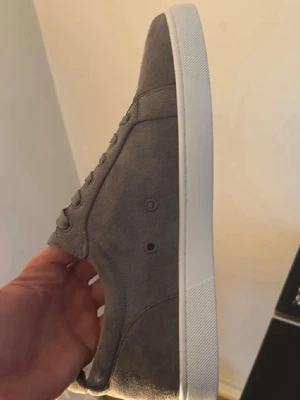 Gråa sneakers från Christian Louboutin - Snygga gråa sneakers från Christian Louboutin i mocka med klassisk röd sula och vita detaljer. Skorna har en låg profil och stilren design med matchande grå skosnören och diskreta sömmar. Perfekta för dig som vill ha något exklusivt och trendigt.