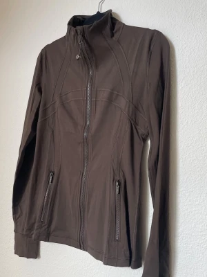 Brun träningsjacka från Lululemon - Snygg brun träningsjacka från Lululemon med hög krage och hel dragkedja framtill. Jackan har två fickor med dragkedja och dekorativa sömmar som ger en sportig look. Tillverkad i ett stretchigt och mjukt material som passar perfekt till träning eller promenader.