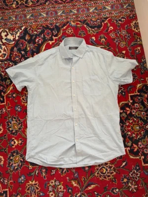 Ljusblå rutig skjorta Dressmann - Kortärmad skjorta från Dressmann i ljusblått med smått rutigt mönster. Klassisk krage med knappar och bröstficka. Perfekt till sommaren och enkel att matcha med jeans eller shorts.