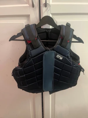 Mörk blå skyddsväst från CRW - Säljer en mörkblå skyddsväst från CRW med vadderade paneler och justerbara axelband. Västen har en sportig design och ger bra skydd vid ridning eller annan sport. Sidorna har snörning för att justera passformen. Perfekt för dig som vill ha extra säkerhet.