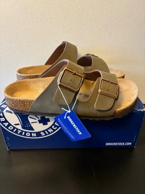 Birkenstock Arizona storlek 41 - Säljer ett par Birkenstock Arizona sandaler i olivgrönt mockaläder med två breda remmar och justerbara metallspännen. Fotbädden är i mjukt naturfärgat läder och sulan är i kork. Klassisk och tidlös modell som är superbekväm.