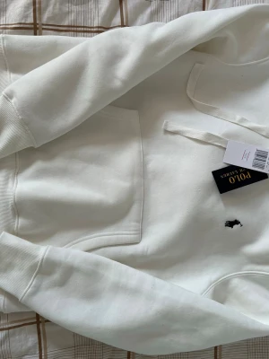 Vit hoodie från Polo Ralph Lauren - Snygg vit hoodie från Polo Ralph Lauren med klassisk broderad logga på bröstet. Hoodien har huva med dragsko och en stor magficka. Tillverkad i mjukt material som känns skönt mot huden. Perfekt för en avslappnad och stilren look.