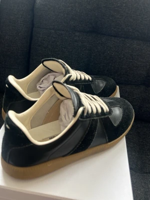 Svarta masion marginella - Snygga svarta Masion marginella  med kontrasterande vita snören och beige innersida. Skorna har en klassisk låg modell med ovandel i både läder och mocka samt en gummisula i ljusbrunt. Perfekta för en stilren och trendig look. Lite använda men inte mycket, bra skick där man får med allt, låda, väska till skorna och de ni ser på bilden 