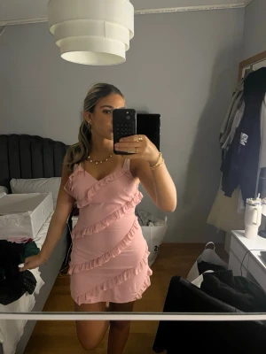 Rosa volangklänning med smala axelband - Söt rosa kortklänning med volangdetaljer och smala axelband. Klänningen har en omlottkänsla och är perfekt för sommarens alla tillfällen. Tillverkad i ett lätt material som ger en bekväm passform.