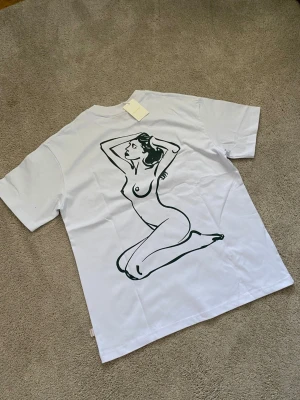 Vit t-shirt Aime Leon Dore med konstmotiv - Vit t-shirt från Aime Leon Dore med svart konstnärlig illustration av en kvinna på ryggen och liten svart text på bröstet. Klassisk passform och rund halsringning. Perfekt för dig som gillar stilrena plagg med unik design.