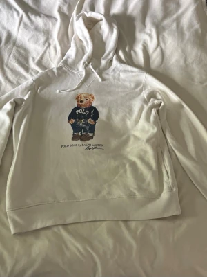 Vit hoodie Polo Bear Ralph Lauren - Säljer en vit hoodie från Ralph Lauren med den ikoniska Polo Bear-trycket på bröstet. Hoodien har huva med dragsko och ribbade muddar. Perfekt för dig som gillar klassisk streetwear med en twist. Materialet är mjukt och skönt, troligen bomullsblandning.