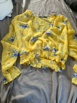 Gul blommig omlottblus - Snygg gul omlottblus med blommigt mönster i lila och blå toner. Blusen har långa ärmar och resår i midjan samt vid ärmsluten för en skön passform. Tillverkad i ett lätt och luftigt tyg som ger en somrig känsla.
