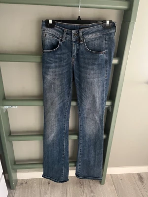 Blå bootcut jeans med F-detalj - Snygga blå bootcut jeans med slitningar och en cool metallisk F-detalj vid fickan. Jeansen har klassisk femficksmodell, normal midja och kontrastsömmar. Perfekta för en avslappnad och trendig look.