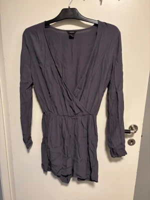 Mörklila långärmad jumpsuit - Snygg grå jumpsuit med långa ärmar och omlott-ringning. Modellen har resår i midjan och korta ben, vilket ger en avslappnad men trendig look. Tillverkad i ett lätt och mjukt tyg som känns skönt mot huden.