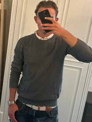 Stickad tröja Dressmann - Snygg mörkgrön stickad tröja från Dressmann med diskret vågmönster. Tröjan har rund halsringning, ribbade muddar och är perfekt att bära över en skjorta för en clean look. Passar till både jeans och chinos.