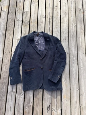 Mörkblå Massimo Dutti kavaj - Stilren mörkblå kavaj i strukturerat tyg med två knappar framtill och bruna detaljer vid fickorna. Kavajen har klassisk krage, knappslå vid ärmslut och rutigt innerfoder. Perfekt för dig som vill ha en dressad men avslappnad look.