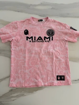 BAPE x Miami t-shirt - Limited edition rosa t-shirt från BAPE x Miami med svart tryck fram och bak. T-shirten har ett subtilt kamouflagemönster, rund hals och korta ärmar. Materialet är mjuk bomull. Storlek L men passar även M. Kom med frågor och prisförslag!☺️