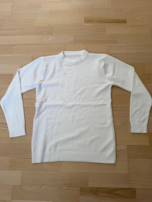 Vit stickad tröja med rund hals - Klassisk vit stickad tröja med rund hals och långa ärmar. Tröjan har ribbade muddar vid ärmslut och nederkant för en clean look. Perfekt basplagg att styla till olika outfits. Materialet ser mjukt och bekvämt ut.