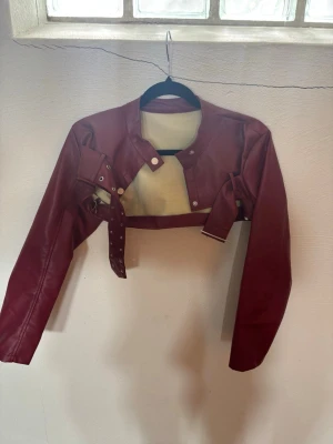 Vinröd croppad skinnjacka - Säljer en vinröd croppad skinnjacka med bälte och silverfärgade detaljer. Jackan har lång ärm, rund halsringning och knappar framtill. Perfekt för dig som vill ha en edgy och trendig look.