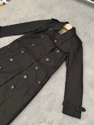 Svart trenchcoat från Burberry - Klassisk svart trenchcoat från Burberry med dubbelknäppning, axelklaffar och bälte vid ärmsluten. Insidan har det ikoniska rutiga Burberry-mönstret. Skriv innan köp!