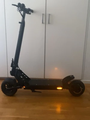 KUKIRIN G2 PRO - Tuff svart elscooter med breda däck och robust ram. Scootern har LED-belysning på sidorna och en stabil plattform för att stå på. Perfekt för att ta sig fram snabbt och smidigt i stan. Handtagen är ergonomiska och designen är modern.
