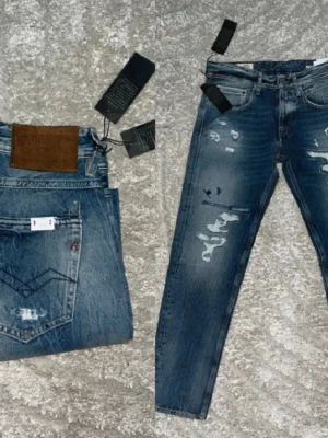 Blå slitna jeans med raka ben - Säljer ett par blå jeans med raka ben och distressed detaljer. Jeansen har flera slitningar och lagade partier framtill för en trendig, avslappnad look. Klassisk femficksmodell med normal midja. Materialet är denim i bomull.