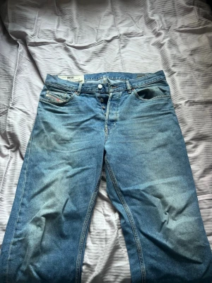 Blå raka jeans från Diesel - Säljer ett par klassiska blå jeans från Diesel med raka ben och knappgylf. Jeansen har fem fickor och snygga slitningar för en avslappnad look. Tillverkade i denim med tydliga Diesel-detaljer på fickan och insidan av linningen. De har ett litet hål i bakfickan, syns knappt 