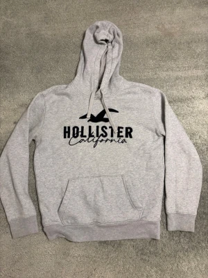 Grå Hollister hoodie med tryck - Snygg ljusgrå hoodie från Hollister med svart logga och texten 'California' på bröstet. Klassisk känguruficka framtill, ribbade muddar och justerbar dragsko i huvan. Perfekt för en avslappnad och trendig stil.