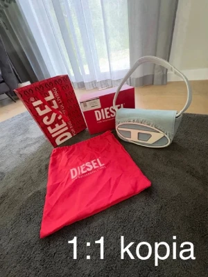 Ljusblå diesel handväska - Trendig ljusblå handväska i denim med vitt handtag och stor vit logodetalj framtill. Väskan har fransad kant upptill och en unik oval form. Perfekt accessoar för dig som gillar statementväskor och streetstyle.