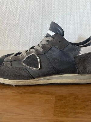 Gråa sneakers i mocka och läder - Snygga sneakers från Phillippe Model i grå mocka och läder med vita detaljer och grå snörning. Skorna har en låg profil och en klassisk sula i beige. Tåpartiet är i textil och designen är stilren med markerade sömmar på sidan.