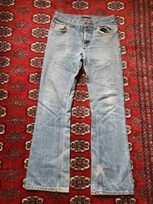 Ljusblå raka jeans från Nudie Jeans - Säljer ett par ljusblå jeans från Nudie Jeans med rak passform och klassisk femficksdesign. Jeansen har orangea sömmar och är tillverkade i denim. Perfekta för dig som gillar en avslappnad och tidlös stil. De är low flare i model.