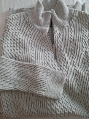 Ljusgrå kabelstickad tröja med halvzip - Supersnygg ljusgrå kabelstickad tröja med halvzip och hög krage. Tröjan har ett klassiskt flätmönster och ribbade muddar vid ärmslut och nederkant. Perfekt för dig som gillar en stilren och mysig look.