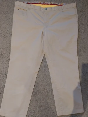 Beiga chinos med färgglad insida - Snygga beiga chinos med rak passform och bälteshällor. Insidan av linningen har ett färgglatt mönster i gult och rött som ger en unik touch. Perfekta för dig som vill ha en clean look med en twist. Stängs med knapp och dragkedja.