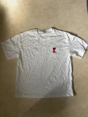 Vit t-shirt med rött hjärta - Säljer en vit t-shirt i bomull med korta ärmar och en broderad detalj på bröstet – ett rött hjärta ovanför bokstaven A. Enkel och clean design som passar till det mesta. Perfekt för dig som gillar stilrena plagg med en twist. Storlek L