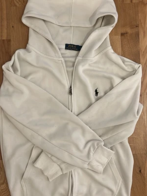 Vit hoodie från Polo Ralph Lauren - Snygg vit hoodie från Polo Ralph Lauren med dragkedja och klassisk logga broderad på bröstet. Hoodien har huva, ribbade muddar och fickor framtill. Perfekt för en chill och stilren look.