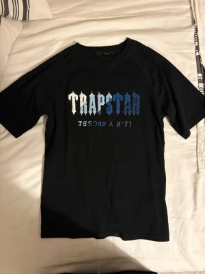 Svart Trapstar t-shirt med broderad text - Säljer en svart t-shirt från Trapstar med broderad text i vitt och blått på bröstet. T-shirten har rund hals och korta ärmar. Materialet känns mjukt och skönt, perfekt för en avslappnad streetwear-look.