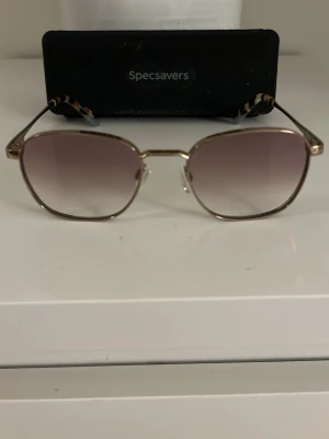 Specsavers solglasögon guld/brun - Snygga solglasögon från Specsavers med guldfärgad metallbåge och fyrkantig form. Glasen är bruna med en snygg toning och skalmarna har ett diskret mönster. Perfekta för soliga dagar och ger en cool vibe till din outfit.