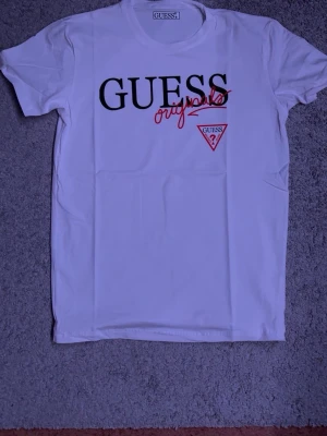 Vit GUESS Originals t-shirt - Snygg vit t-shirt från GUESS Originals med svart och rött tryck framtill. Klassisk passform med korta ärmar och rund halsringning. Stort GUESS-tryck och logga på bröstet ger en clean och trendig look. Perfekt till jeans eller shorts.