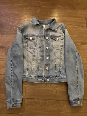 Ljusblå klassisk jeansjacka - Ljusblå jeansjacka i klassisk modell med raka linjer och silverfärgade knappar. Jackan har två bröstfickor med lock och knapp samt knapp ”dragkedja” framtill. Tillverkad i denim med lätt tvättad look och krage.
