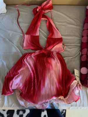 Plisserad rosa och röd halterneckklänning - Säljer en kort plisserad klänning i rosa och rött med halterneck och knyt i nacken. Klänningen har ett omlottliknande fall och är tillverkad i ett lätt, glansigt material som ger en snygg effekt. Perfekt för dig som vill sticka ut med färg och form!