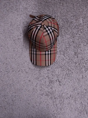 Burberry keps - Snygg beige keps med klassiskt rutmönster i svart, vitt och rött. Kepsen har böjd skärm och justerbar rem bak. Perfekt accessoar för en trendig streetwear-look.