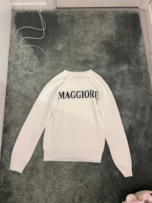 Maggiore - 160cm Använd en gång helt ny. .Säljer en vit stickad tröja med rund hals och ribbade muddar. På ryggen står det 'MAGGIORE' i svart text. Tröjan har en clean look och är perfekt för dig som gillar enkel men snygg stil.