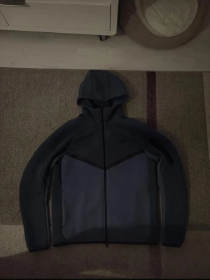 Mörkblå hoodie med dragkedja - Snygg mörkblå hoodie med hel dragkedja och huva. Hoodien har en stilren design med svarta detaljer och två fickor framtill. Perfekt för dig som gillar en enkel men modern look. Materialet känns mjukt och bekvämt.