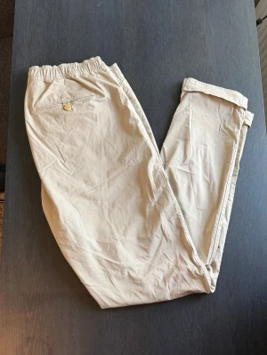 Beige mjukisbyxor med dragsko - Säljer ett par beige mjukisbyxor med resår i midjan och dragsko för skön passform. Byxorna har fickor på sidorna och en bakficka med knapp. Tillverkade i ett mjukt och lätt material, perfekta för chill dagar.