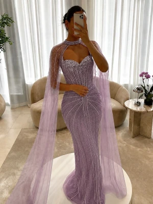 Lång lila glittrig balklänning - Magisk lång lila klänning med glittriga detaljer och transparent cape. Klänningen har hjärtformad urringning, figurnära passform och är perfekt för bal eller fest. Materialet är glittrigt och skimrande, vilket ger en lyxig känsla.